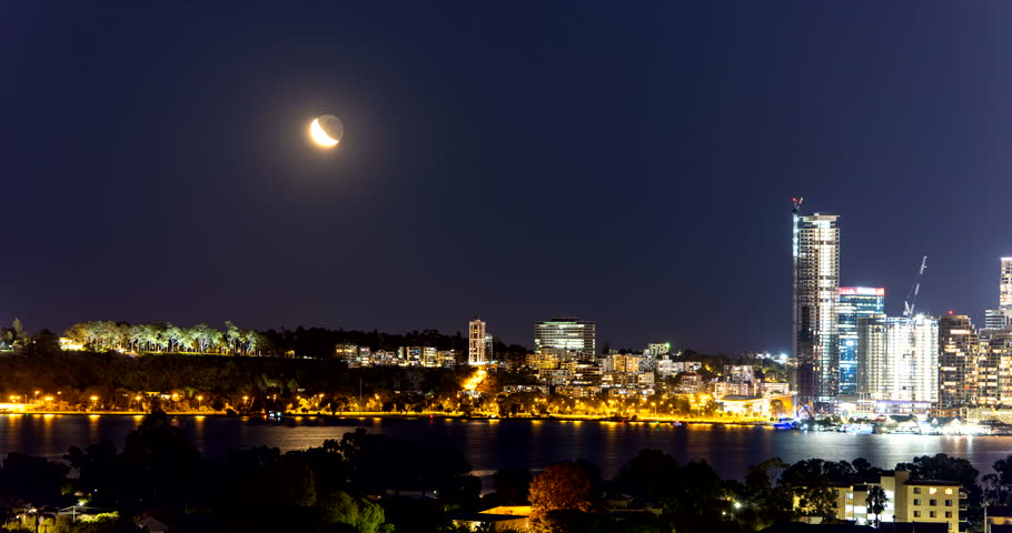 A serene night view of Perth, featuring a crescent moon over the Swan River. Warm city lights reflect on the water, highlighting the city modern skyscrapers, tree-lined parks, and tranquil atmosphere