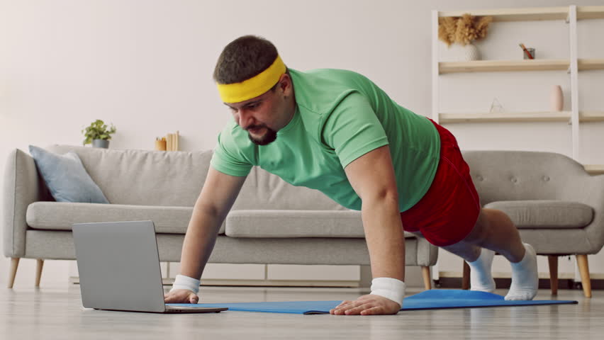 A man focused on his workout routine is seen doing push-ups at home. He wears a green shirt, red shorts, and headband, following a fitness routine displayed on his laptop.