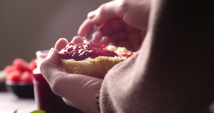 A close-up of a spoon spreading vibrant raspberry or strawberry jam onto freshly torn bread. Rich jam glistens under soft lighting, with a jar of jam and fresh raspberries blurred in the background.