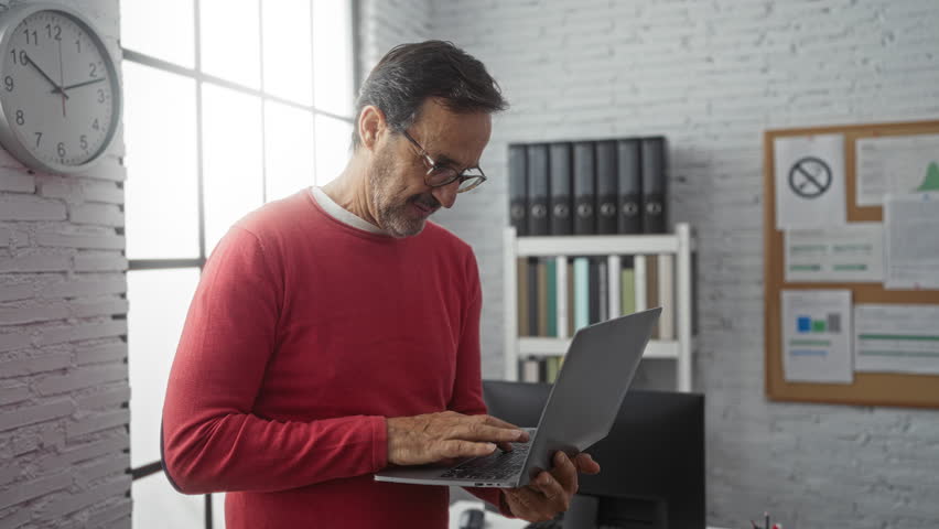 Mature hispanic man in red sweater uses laptop in modern office with clock and shelves. - Powered by Shutterstock - Get 15% off with code: PIKWIZARD15