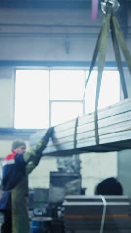 A factory worker uses a crane to lift heavy metal beams in an industrial workspace. The activity showcases teamwork and machinery in action during a busy workday.