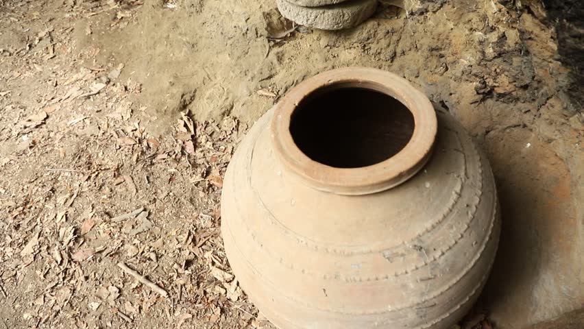 A rustic, dimly lit scene inside a cave or rock shelter. Old pottery jars, a wooden barrel, a woven basket, and a stone millstone are scattered on the uneven floor and shelves. 