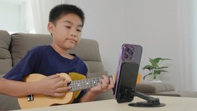 Asian kid boy playing ukulele while live-streaming his music lesson using a smartphone. The mounted phone stand and relaxed living room scene showcase modern learning and creative activities at home. - Powered by Shutterstock - Get 15% off with code: PIKWIZARD15