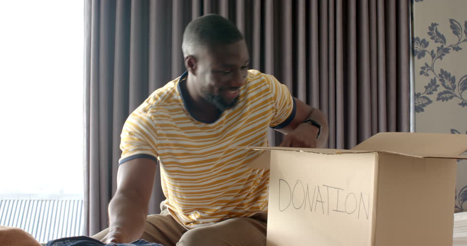 Packing clothes into donation box, African American man smiling and organizing items. Charity, volunteering, helping, giving, community, unaltered, lifestyle