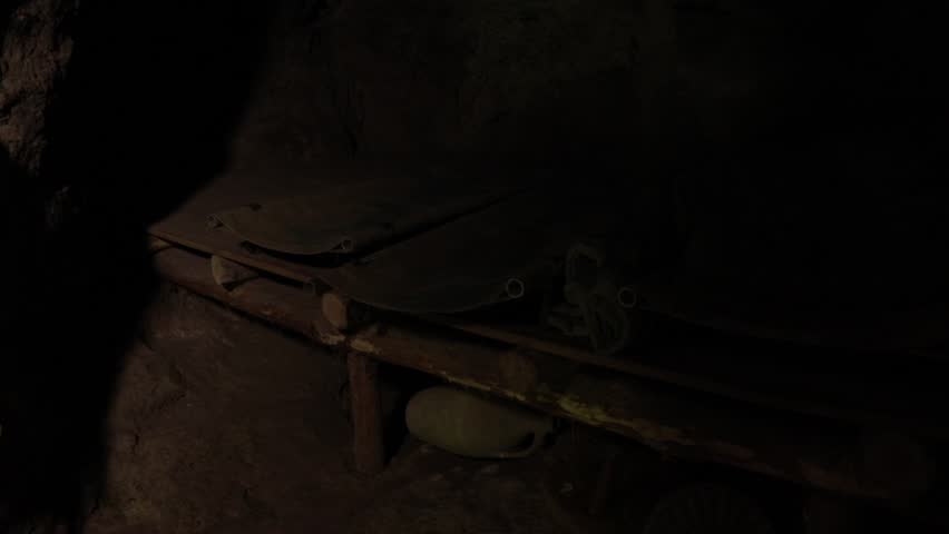 A dim, cave-like space shows rudimentary cots made of wooden logs. Two cots are canvas and a sleeping bag sits on one. An old jar rests beneath. Scene suggests a rustic shelter.