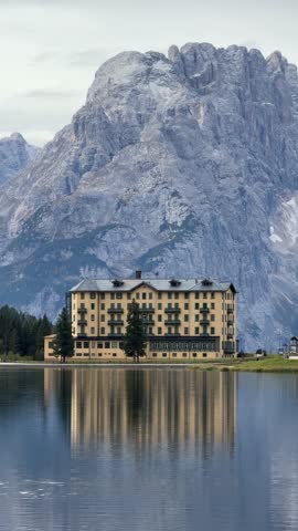 A large building is reflected in the water. The building is surrounded by mountains. The mountains are covered in snow