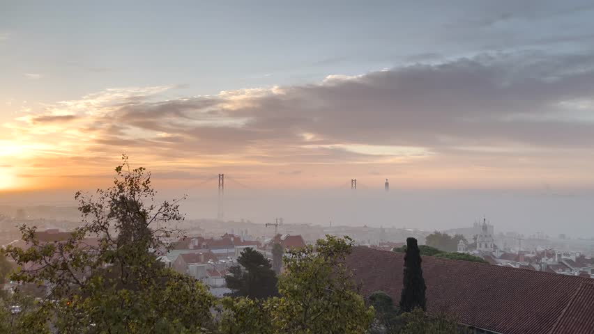 Morning mist shrouds the 25 de Abril Bridge and Tagus River in Lisbon, Portugal, creating a dreamy and atmospheric view of the cityscape. A blend of nature and urban beauty.