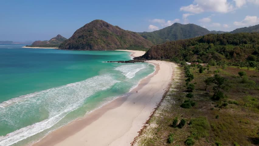 4K drone video of a couple walking along Selong Belanak Beach in Lombok, Indonesia. The long white sand beach has white waves rolling in from the clear blue ocean with mountains surrounding the beach