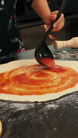 Woman spreading tomato sauce on pizza dough with ladle, making traditional italian pizza. Female pouring tomato sauce over pizza. Process of making homemade pizza. Vertical video