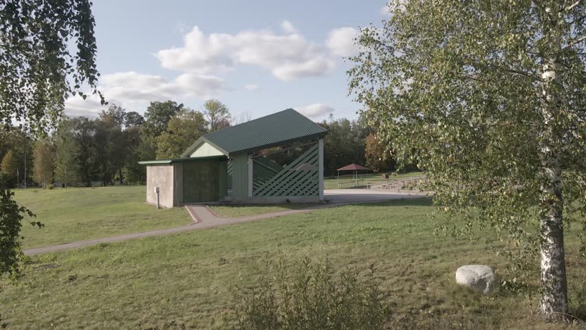 Open seating area with wooden benches, picnic tables under red roofed shelters, grassy field, trees with autumn hues, and a partly cloudy sky.