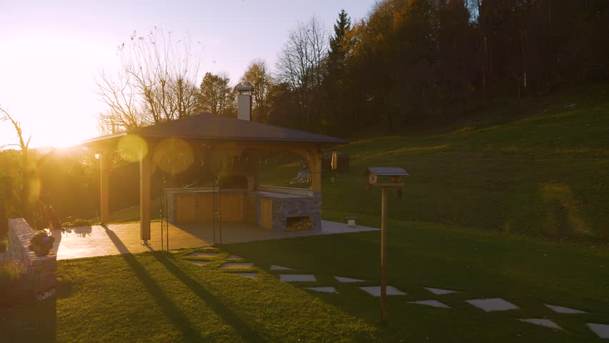 LENS FLARE: Backyard pavilion with outdoor kitchen at sunset, surrounded by scenic hills and lush greenery in autumn shades. Golden sunlight casts long shadows on modern garden overlooking the valley