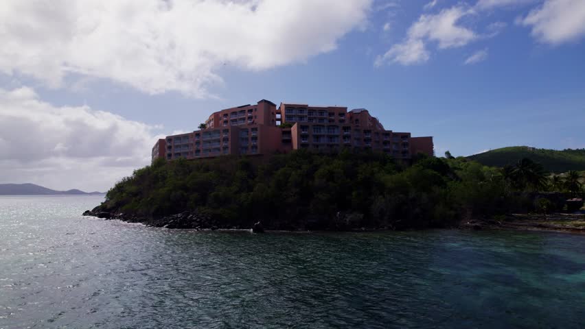 Aerial pullback over water of St. Thomas, USVI, highlighting oceanfront cliffs and vibrant tropical surroundings, backlit hotel building