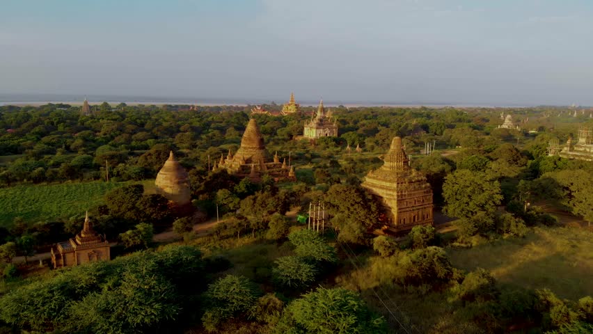 Majestic temples rise from the lush landscape of Bagan, Myanmar as golden sunlight casts shadows on ancient structures. Witness the serene beauty of this historic site awakening to a new day.