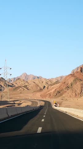 Vertical front view of car driving motion by empty asphalt highway in the desert with road surface marking lines. Sand mountains on the roadside and the hot sun in the blue sky