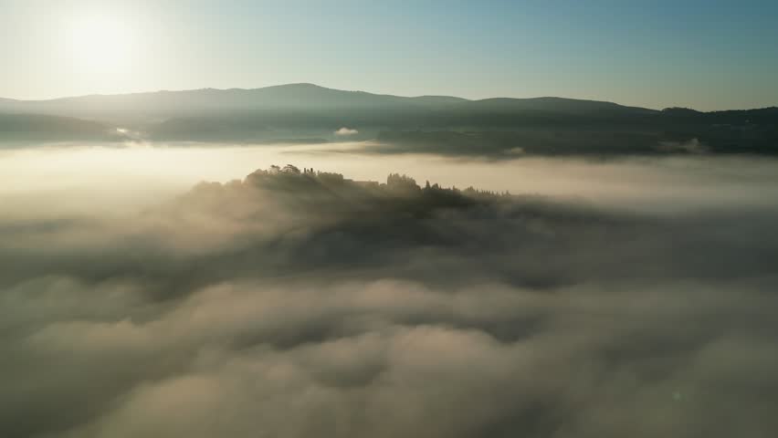 Aerial drone footage of a serene Tuscany sunrise, with golden rays piercing the mist over lush hills and treetops, creating a dreamlike spring landscape.