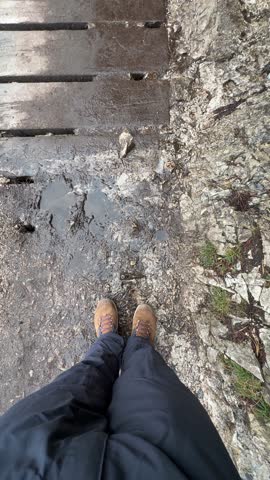 A person is standing on a bridge with their feet in the water. The bridge is old and has a lot of cracks. The water is murky and dirty. The person is wearing boots and is looking down at the water