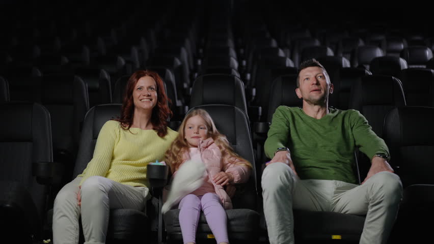 A family of three arrives at an empty cinema hall and settles into the first row. The mother, wearing a yellow sweater, smiles as she sits down, while the father, dressed in a green sweater, helps