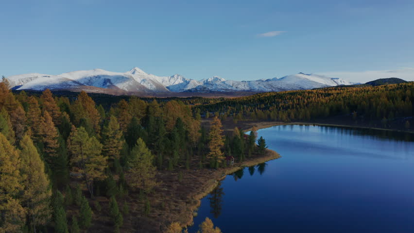 Pan left experience a stunning aerial view of Mountain Altai captured by a drone during late autumn, showcasing vibrant foliage and snow-capped peaks mirrored in a serene lake.