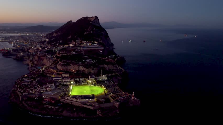 Aerial view of Gibraltar at sunset. Street lights on. Europa Sports Complex Stadium with night light on. 
 Bay of Gibraltar. City of Gibraltar and La Linea, in background. Track left. Travel destinati