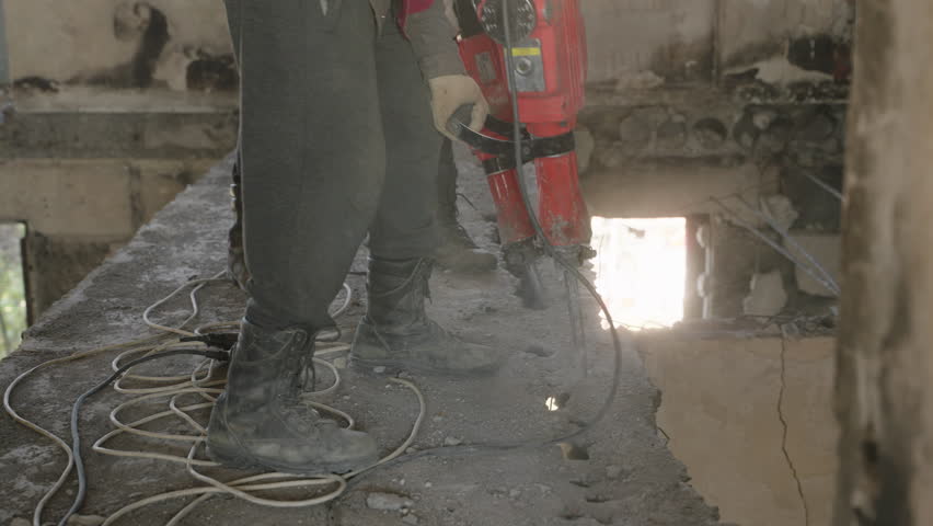 Construction workers use jackhammers to demolish damaged walls, ceilings in war-affected apartment. Dust-filled air, debris from concrete in renovation process. Laborers in protective gear working.