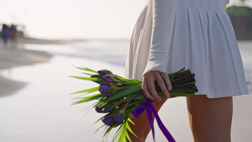 Female legs going with bouquet of purple flowers along sandy ocean coast. Slim person