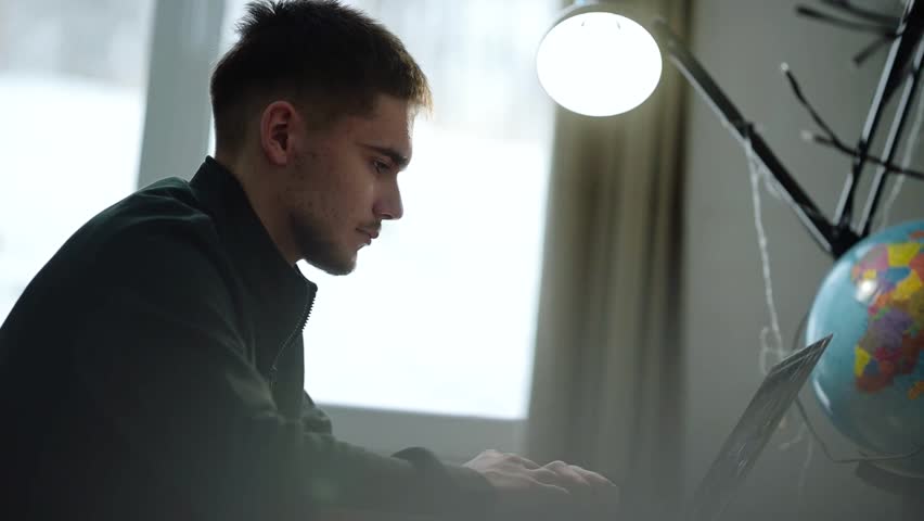 Young man focused on work at a laptop near a globe in a cozy indoor setting during the day