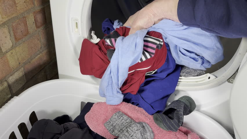 Closeup slow motion shot of a man’s hand pulling a variety of washed and dried clothes from a front loading tumble dryer straight into a laundry basket on the floor.