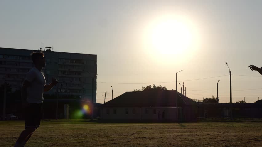 Silhouette of professional footballers kicking soccer ball with their heads on stadium at sunset. Two sportsmen showing tricks with ball while passing it to each other at field. Freestyle football