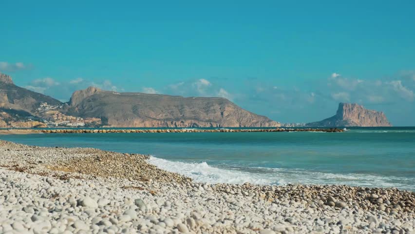 White pebble beach in Altea and beautiful Mediterranean Sea. View from Altea to turquoise Mediterranean sea with rolling waves, Calpe city, Rock of Ifach in sunny day. Altea, Alicante province, Spain