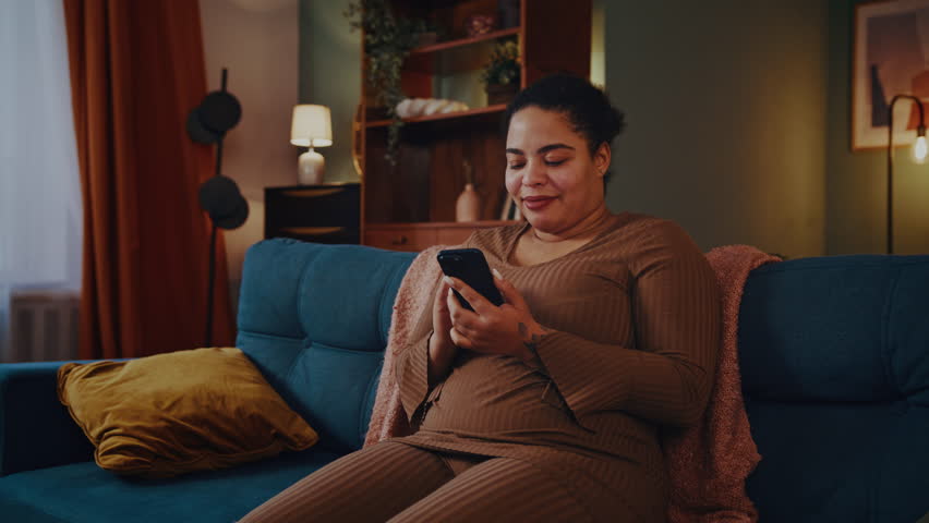 Happy African American woman holds smartphone expressing joy. Plus size female screams Yes making dance movements sitting on sofa in living room