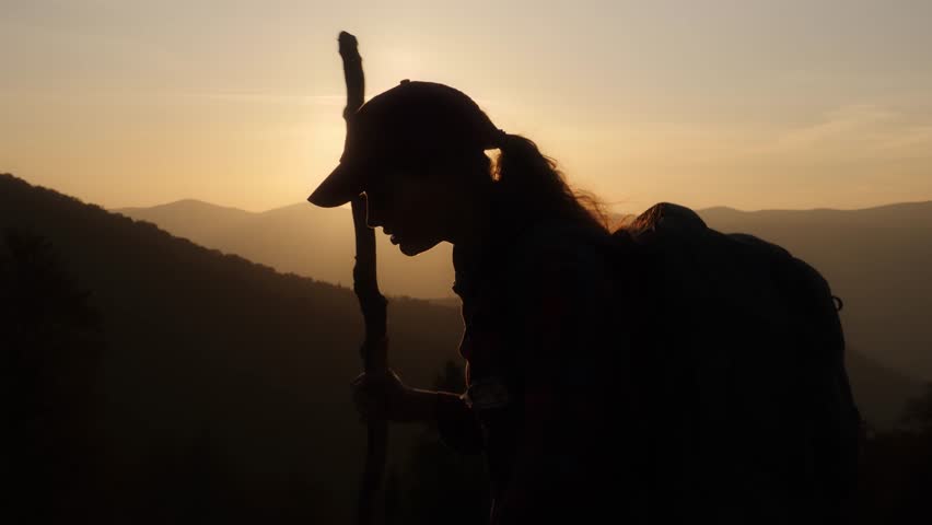 Handheld slow-motion silhouette of a female hiker with a backpack and walking stick, moving against a sunset backdrop in the mountains. Perfect for themes of adventure, nature, and serene travel