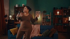Mixed race woman dances singing favourite song against sofa in living room. Happy plus size female uses TV remote control as microphone having fun - Powered by Shutterstock - Get 15% off with code: PIKWIZARD15