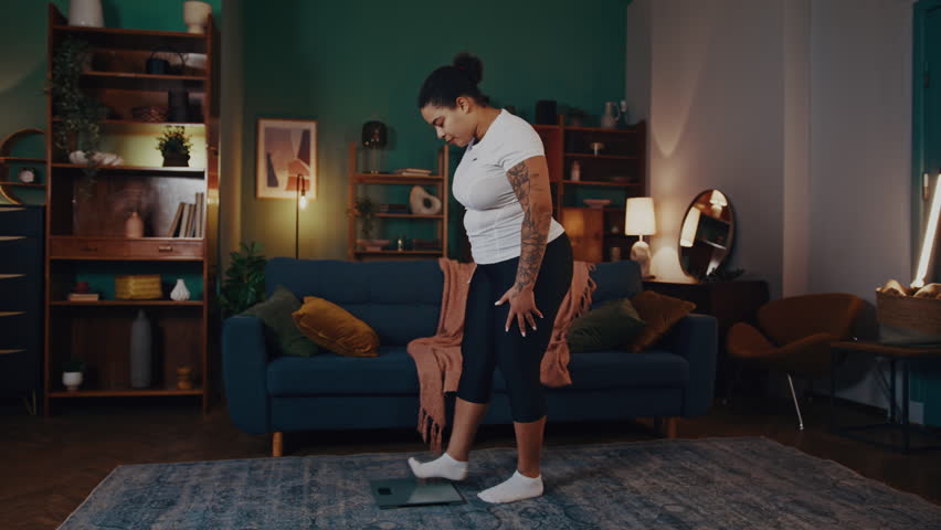 African American woman steps on scales expressing joy with results. Plump female reflects positive emotions due to loss of several kilograms after training