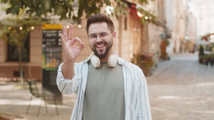 Portrait of smiling young millennial guy looking at camera showing hand ok sign. Caucasian handsome man showing everything fine success gesture body language. Tourist in glasses smiling at camera.