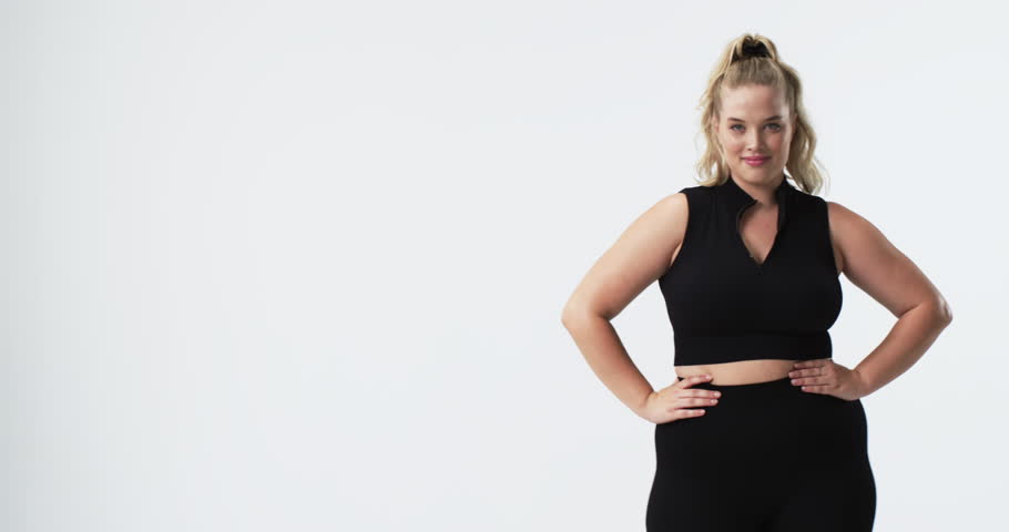 Caucasian plus size woman with light skin, blonde hair, and blue eyes flexing arm and smiling, copy space. She is wearing black workout clothes and standing against plain white background, unaltered