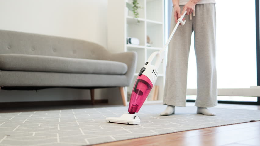 Young Asian woman vacuuming living room carpet with handheld vacuum cleaner. Clean and tidy home environment emphasizing daily household chores and contemporary lifestyle.