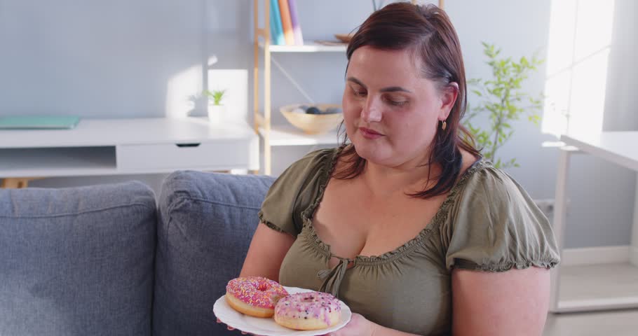 Happy smiling overweight woman enjoying donuts while sitting on sofa at home. Satisfied fat female indulging in sweet treats, showing carefree attitude, love for desserts and unhealthy eating habits.