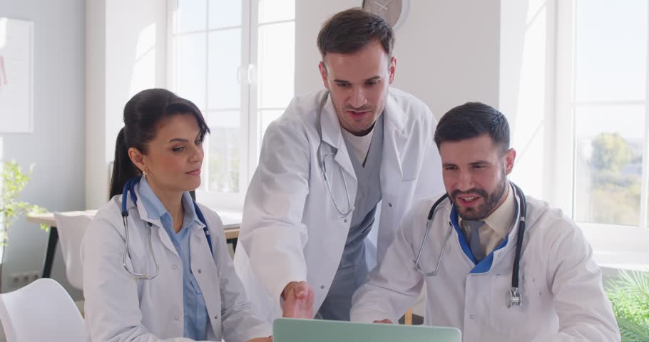 Team three happy smiling doctors in white medical coats working in office, using laptop computer, marking successful development of new effective cure or diagnostic method, giving each other high five
