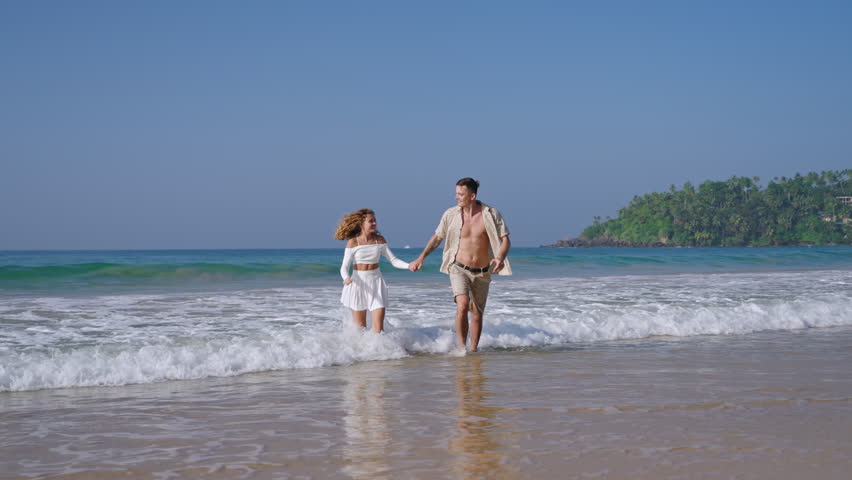 Attractive couple run holding hands in sea surf waves. Tanned man and woman having fun on summer vacation at the sandy beach. Boyfriend, girlfriend enjoying romantic date. Slow motion.
