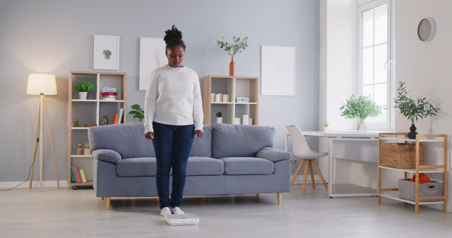 Unsatisfied African american woman doing weight measurement on floor scales, getting bad, not good enough result, feels overweight during regular check, monitoring body mass, loss of weight, slimming 