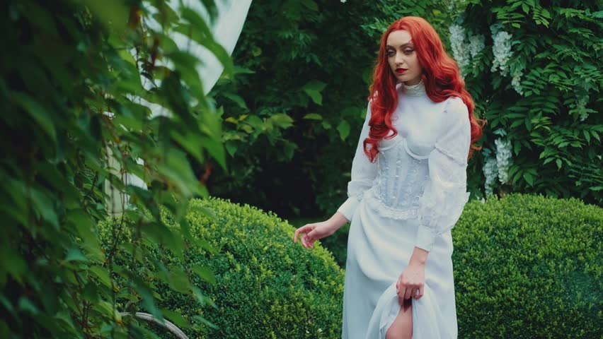 Fantasy red-haired dangerous woman medieval assassin queen with dagger in sheath on leg leather belt. White vintage old style dress. Girl princess warrior. Green summer nature trees. Hand hold knife.