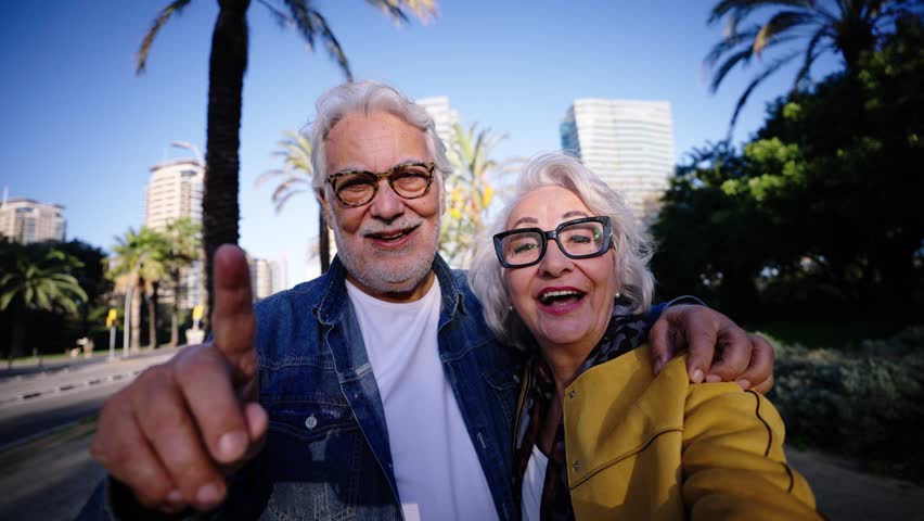 Caucasian smiling pensioners in love couple doing a video call selfie hugging together outdoors. Happy romantic adult people embracing. Grandparents look camera affectionate enjoying retirement time