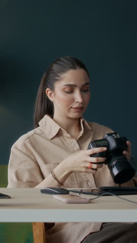 Vertical shot of young female Caucasian photographer checking photos on camera while sitting at desk indoors