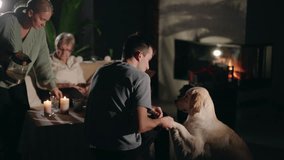 A family enjoys a cozy evening by the fireplace. A woman pours tea into cups, an elderly woman relaxes nearby, and a young man plays affectionately with a Golden Retriever. High quality 4k footage - Powered by Shutterstock - Get 15% off with code: PIKWIZARD15