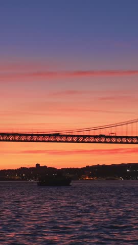 View of 25 de Abril Bridge famous tourist landmark of Lisbon connecting Lisboa and Almada on Setubal Peninsula over Tagus river with passing tourist yacht boat silhouette on sunset. Lisbon, Portugal