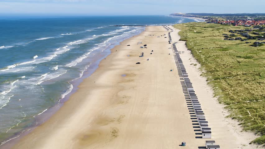 Drone flying sideways over the popular Løkken beach in Denmark. There is a long row of small beach huts, and there are people walkning and running. It is a sunny spring day