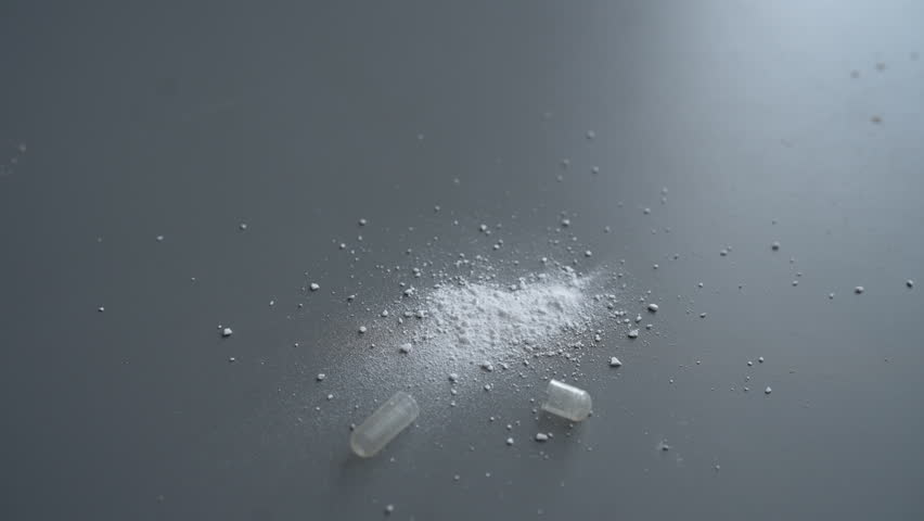 Close-up of white pharmaceutical capsule crashed, releasing fine powdery contents spreading across textured gray surface, revealing granular pharmaceutical substance details, nobody, slow motion.
