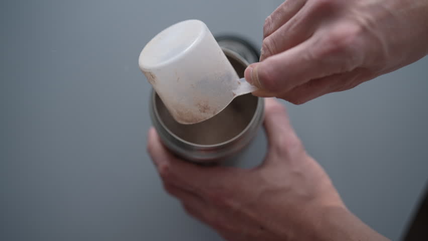Muscular athlete using scoop to pour protein powder into shaker bottle, preparing nutritious post-workout supplement or meal replacement for optimal recovery and muscle gain. Shooting in slow motion.