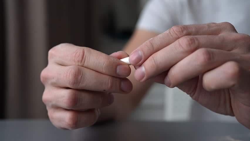 Cropped shot of male hands crushing pharmaceutical capsule, grinding medication into fine white powder on gray surface. Concept of medicine, health, wellness, healthcare, treatment, close-up, slowmo.