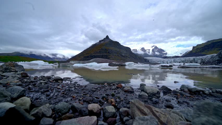 Mountainous area. In the foreground, there are shallow pools of water reflecting the sky. Behind them, there is a small, triangular wooden hut situated on a grassy plain.